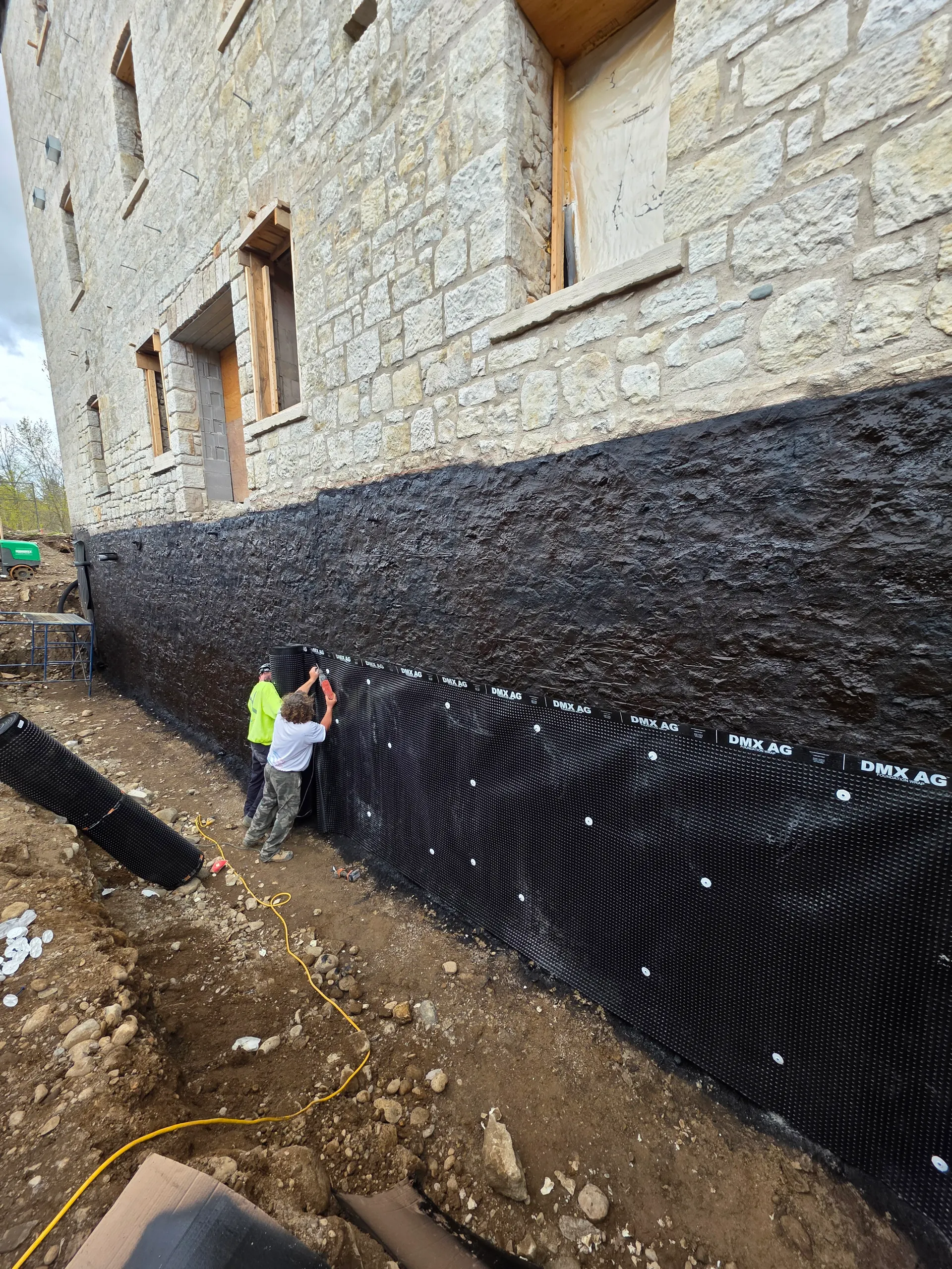 Crew installing a below-grade waterproofing membrane and drainage board on a stone foundation wall