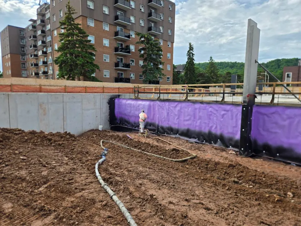 Purple below-grade waterproofing membrane being spray-applied along a foundation wall